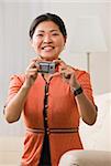 A woman is holding a camera in her hands.  She is smiling at the camera.  Vertically framed shot.