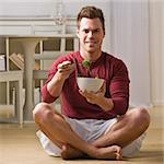 Attractive man smiling at camera, sitting in lotus and eating salad. Square.