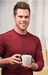 A young man is holding a cup of coffee and smiling at the camera.  Vertically framed shot.