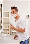 Un homme est raser son visage devant le miroir de salle de bains. Il est souriant à la caméra. Photo encadrée verticalement.