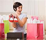 A young woman is surrounded by shopping bags and is holding out a pair of shoes.  She is smiling at the camera.  Square framed shot.