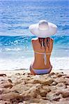 Beautiful brunette with long hair in white hat and swimsuit sits on the sand ocean beach on Bali in Indonesia