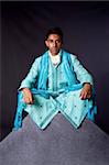 Beautiful authentic Indian hindu man in typical ethnic groom attire sitting in lotus position with legs crossed on top of rock. Bangali male wearing a light blue agua decorated Dhoti with shawl.