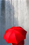 A woman dressed in a red jacket and carrying a red umbrella standing in front of a waterfall. Shot on location at Skogafoss in Iceland.