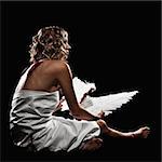 A young, adult female model is sitting and draped in white fabric. She is in profile and looking away from the camera. She is holding wings that she has taken from her back as indicated by bloody back wounds. The shot is in a square format.