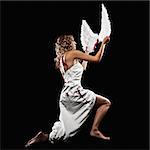 A young, adult female model is kneeling and draped in white fabric. She is in profile and looking away from the camera. She is holding wings that she has taken from her back as indicated by bloody back wounds. The shot is in a square format.