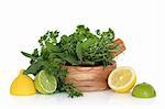 Lemon and lime fruit with mixed herb leaf selection in an olive wood mortar with pestle, over white background.