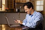 Man sitting at table smiling with cell phone and laptop. Horizontally framed shot.