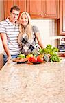 Young couple smile into the camera behind a kitchen counter.  The counter is displaying a variety of fresh vegetables. Vertical shot.