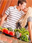 Low angle, tilted view of young couple kissing behind a kitchen counter holding fresh vegetables. Vertical shot.
