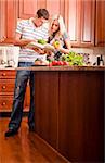 Young couple examine a recipe book in the kitchen. The counter is full of fresh vegetables. Vertical shot.