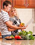 Young woman looks back affectionately at a young man as he reads a recipe book. A kitchen counter holds a variety of fresh vegetables in the foreground. Vertical shot.