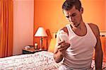 Man sits on his bed and looks pensively at a bottle of pills. Horizontal format.