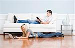 Man reads on a couch while woman stretches out on the floor with her laptop. Horizontal format.