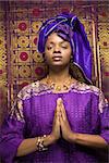 Portrait of an African American woman wearing traditional African clothing in front of a patterned wall and holding her hands in a prayer position. Vertical format.