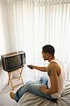 High angle view of a young man sitting on the edge of a bed and turning on the television. Vertical shot.