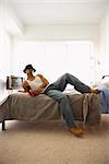 A young man lying on a bed. He is dressed in casual clothing and wearing a hat. Vertical shot.