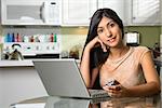 Portrait of a young woman sitting at the kitchen table with a laptop and a cell phone. Horizontal shot.