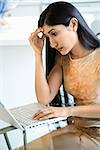 A young woman sitting at a glass kitchen table using a laptop. She has a worried expression on her face. Vertical shot.