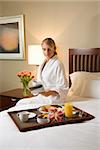 Caucasian woman wearing a bathrobe sits on a hotel bed with tray of breakfast. She is reading a newspaper and holding a coffee cup. Vertical format.