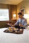 Caucasian businessman enjoys his breakfast while reading the morning paper. He is sitting on the bed and holding a cup of coffee. Vertical shot.