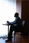 African-American businessman sitting down at a small cafe table next to a curtained window. He has a coffee cup in hand. Vertical shot.