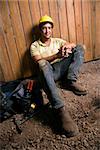 Male Caucasian construction worker sitting next to a tool bag.  He is wearing a yellow hardhat. Vertical shot.