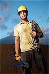 Attractive male construction worker in a yellow hardhat smiles into the camera. He holds a heavy chain on his shoulder and bolt cutters in his hand. Vertical shot.