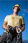 Attractive male construction worker wearing a yellow hardhat and work gloves looks down at the camera while holding a heavy chain and hook. Vertical shot
