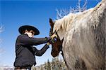 Attractive young man grooming his horse with a snowy mountain landscape in the background. Horizontal shot.