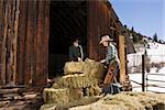 Attractive young man and woman lifting hay bales on a farm in front of a barn. Horizontal shot.