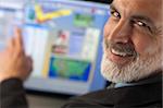 Close-up portrait of a businessman smiling over his shoulder at the camera while pointing to a computer monitor. Horizontal shot.