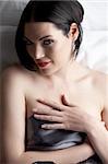 Sexy naked young caucasian adult woman with red lips, short black hair and a pierced eyebrow, covered in a dark satin sheet and sitting on a bed