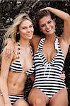 Portrait of two attractive young women wearing swimsuits with the same striped pattern and leaning against a large rock. They have their arms around each other and are smiling at the camera. Vertical shot.