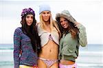 Three attractive young women in bikinis are standing on the beach.  They are also wearing sweaters, a coat and knit caps. Horizontal shot.