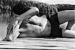 Young man in swimming trunks is lying against lake / black and white picture