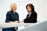 Two businesswomen sitting at office desk looking and talking over papers.