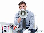Furious businessman yelling through a megaphone sitting at his desk in the office
