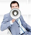 Furious young businessman yelling through a megaphone sitting on his desk