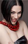Sexy naked young caucasian adult woman with red lips, short black hair and a pierced eyebrow, covered in a dark satin sheet and wearing a red coral necklace