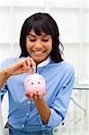 Enthusiastic ethnic businesswoman saving money in a piggybank in the office