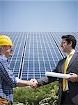 Portrait of mid adult italian male engineer holding blueprints and shaking hands to manual worker in solar power station. Vertical shape, side view. Copy space