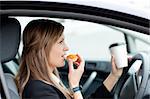 Charming businesswoman eating and holding a drinking cup while driving to work