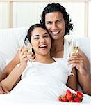 Happy young lovers drinking Champagne with strawberries on the bed