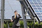 Portrait of mid adult italian male engineer holding blueprints in solar power station and talking on mobile phone. Horizontal shape, front view. Copy space