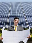 Portrait of mid adult italian male engineer reading blueprints in solar power station and smiling. Vertical shape, front view. Copy space
