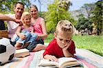 Concentrated blond boy reading while having a picnic with his family in a park