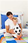 Cute little boy and his father playing with a soccer ball sitting on bed