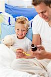 Smiling father giving cough syrup to his sick son sitting on bed