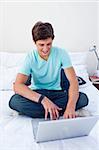 A smiling teenage guy sitting on his bed using a laptop
